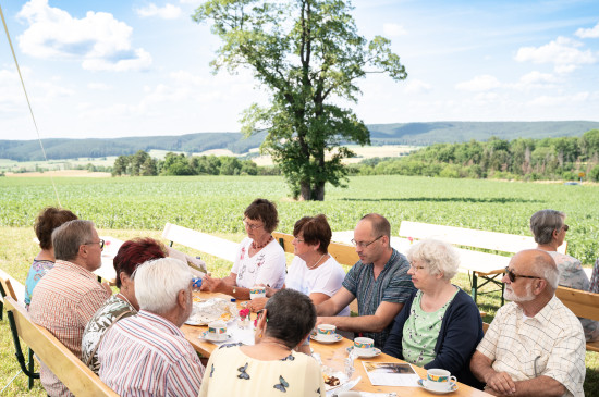 Menschen sitzen an einem langen Tisch beisammen, der Blick geht ins Grüne in die Weite